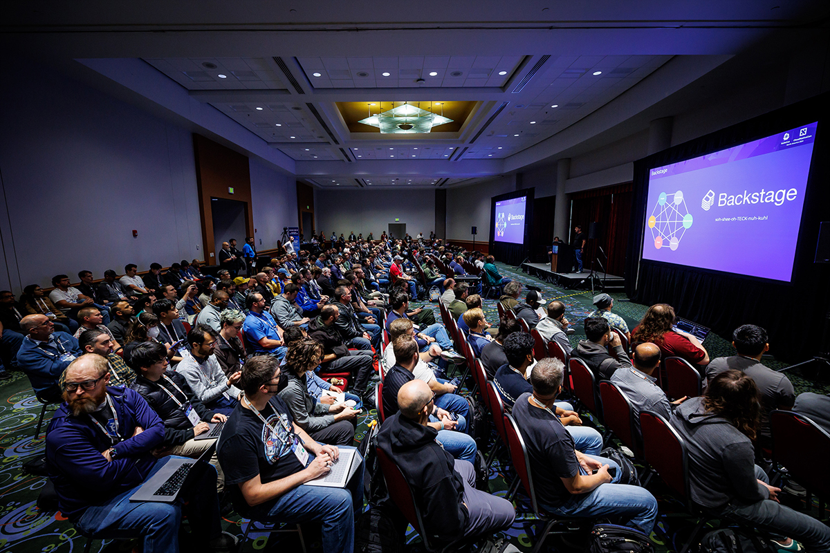 A packed ballroom with Backstage displayed on two large screens